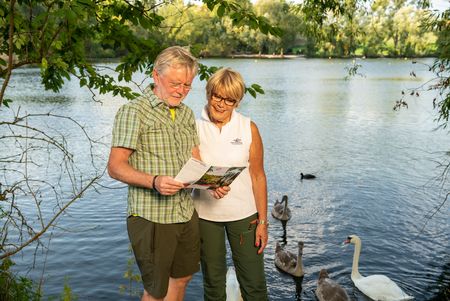 Frau und Mann lesen Wanderflyer des neanderland STEIGs am Ufer mit Schwänen am Abtskücher Teich in Heiligenhaus